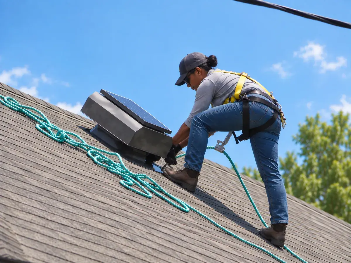 Roofing professional in a safety harness installing an Attic Breeze solar attic fan on a shingle roof